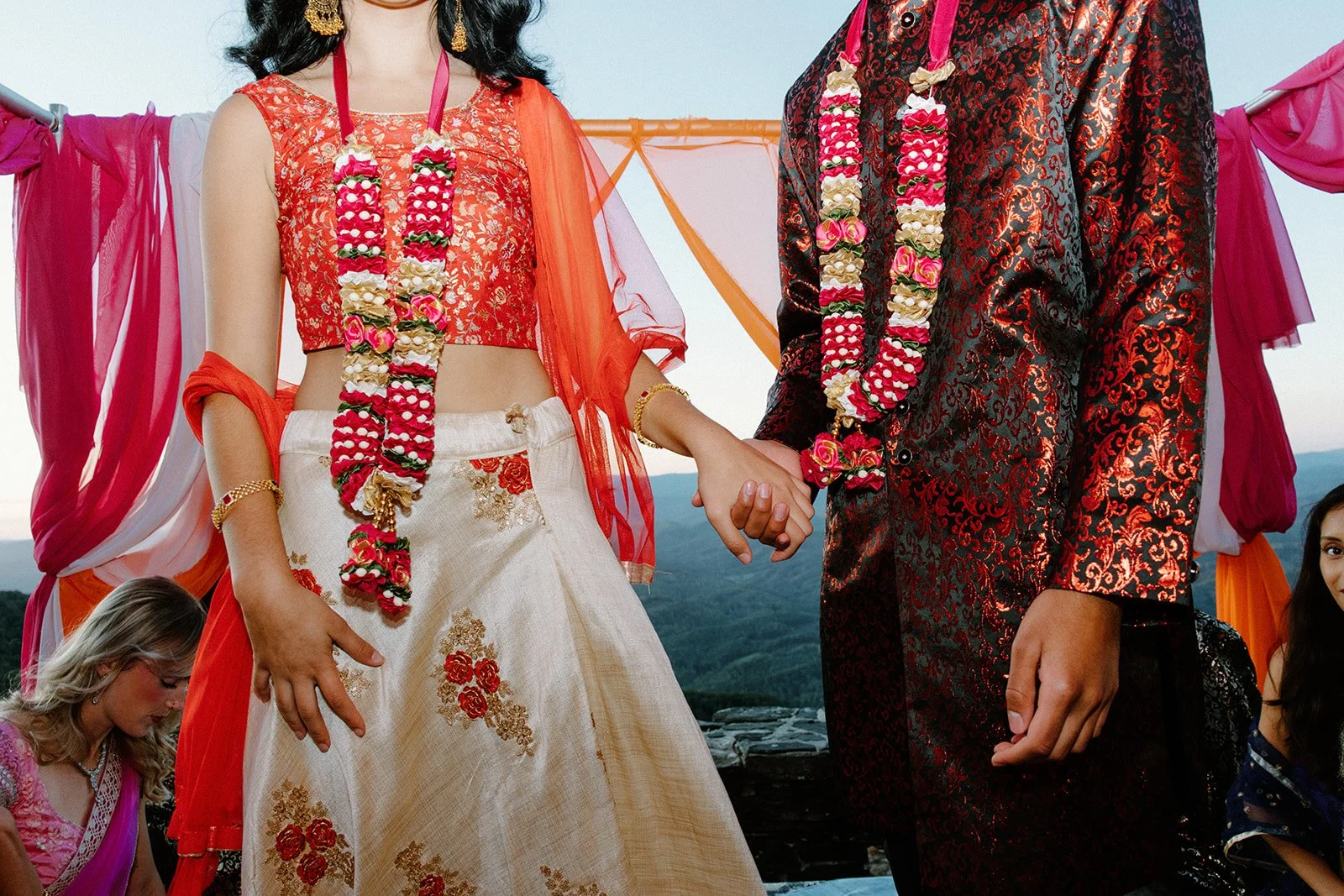 Cropped view of couple holding hands, wearing red-and-white flower garlands and formal South Asian wedding attire.