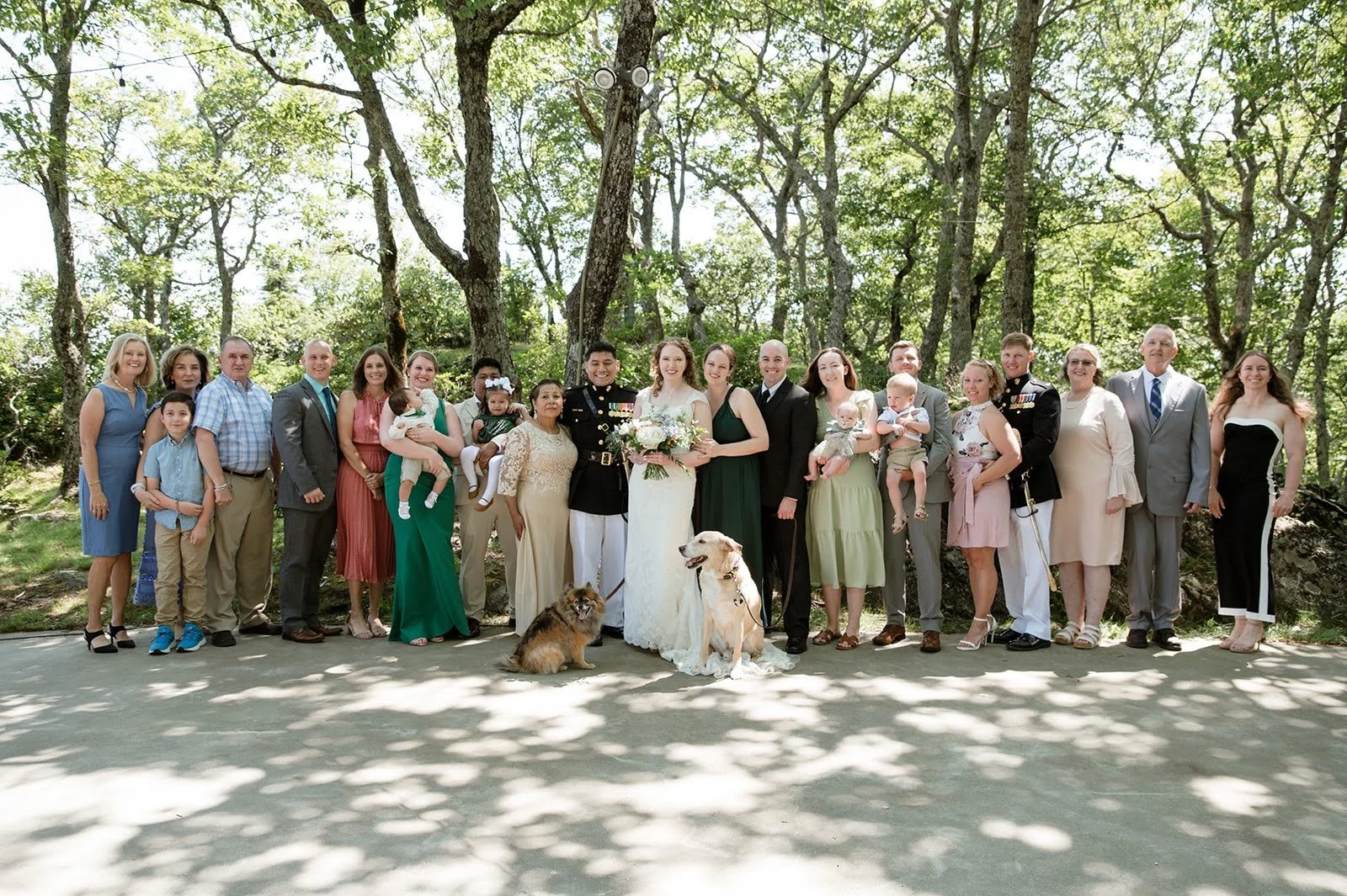 Large family wedding portrait with the bride, military groom, guests, children, and dogs in a wooded venue.