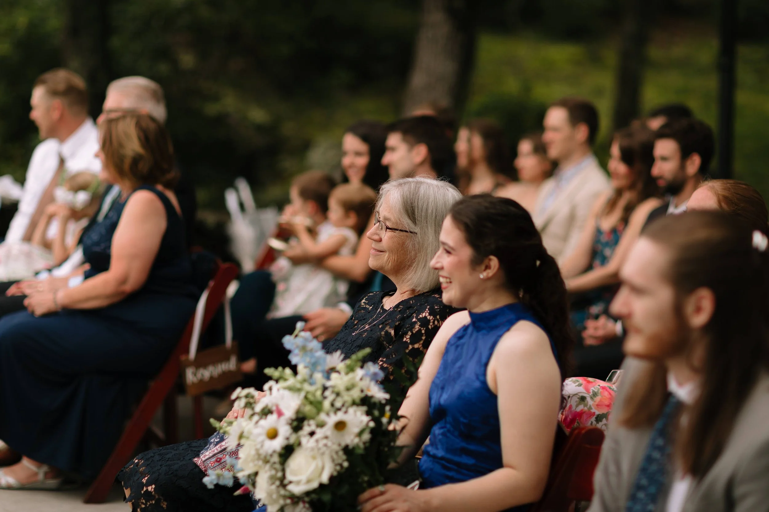 Wedding guests seated outdoors during a ceremony with a white floral bouquet in the foreground.
