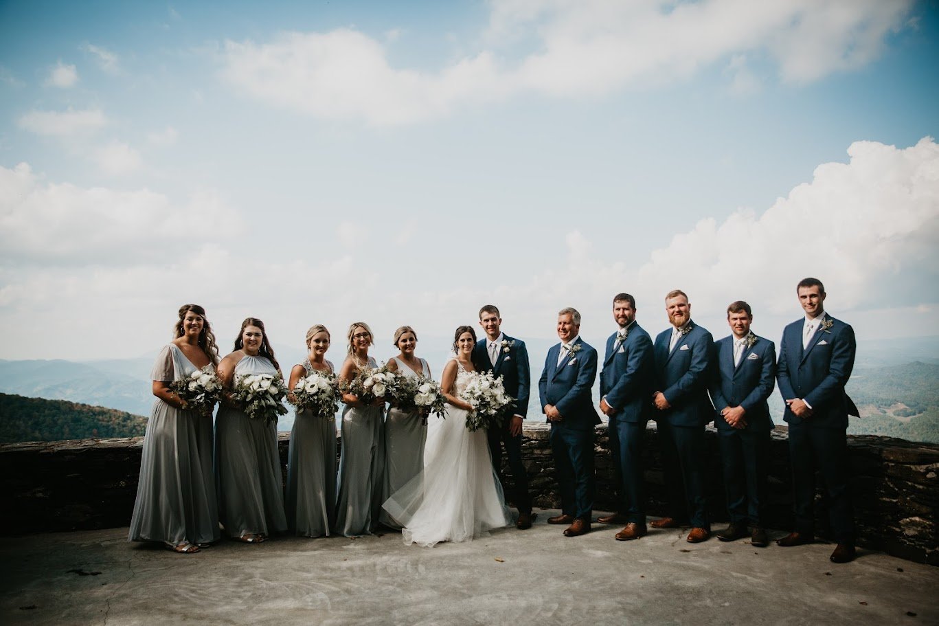 Bride and groom posing with their wedding party at a mountain overlook with gray bridesmaid dresses and navy suits.