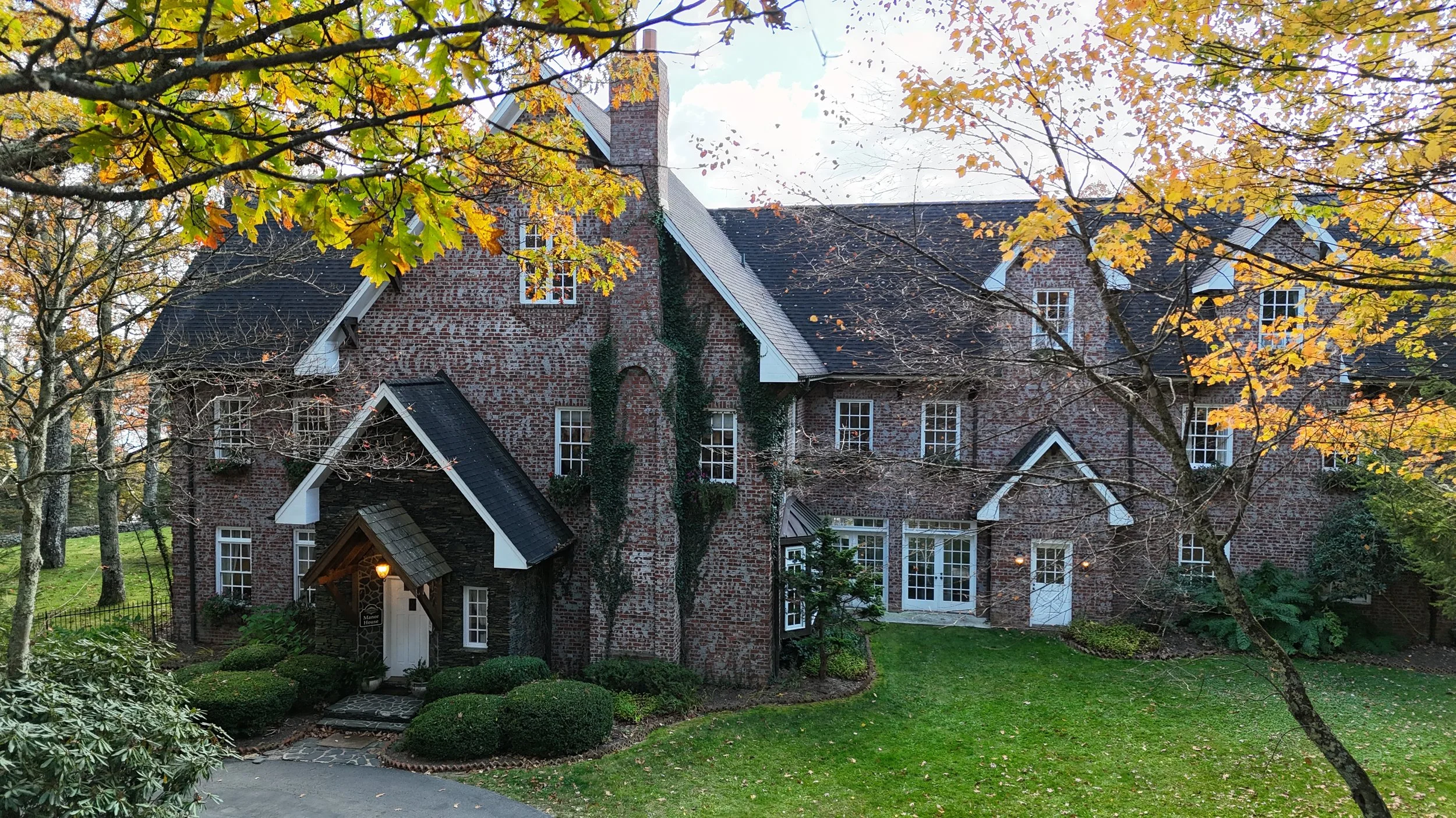 Brick manor exterior with steep gables and ivy, framed by fall leaves, with a green lawn and front entry path.
