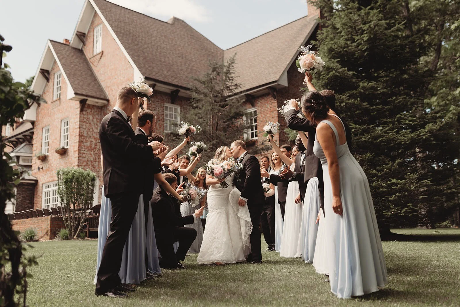 Wedding party cheering as the bride and groom kiss on the lawn in front of a brick estate venue