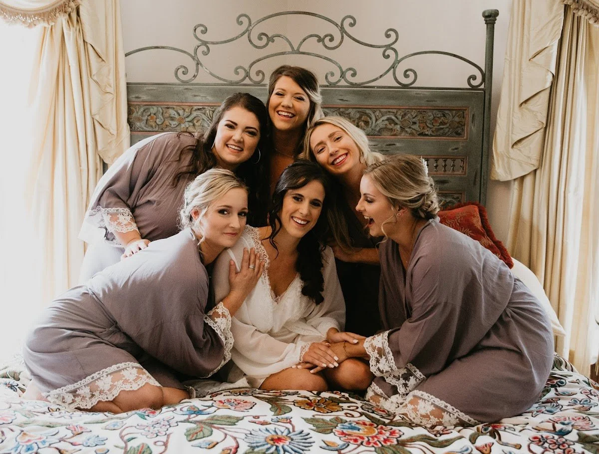 Bride and bridesmaids sitting together on a bed in robes for a relaxed getting-ready portrait.