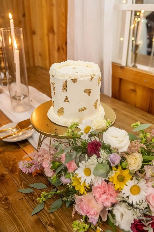 Small white cake with gold leaf accents on a stand beside a colorful floral arrangement on a wooden table.