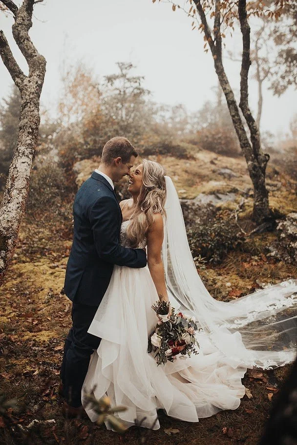 Bride and groom touching foreheads in a fall woodland setting with the bride’s long veil trailing.