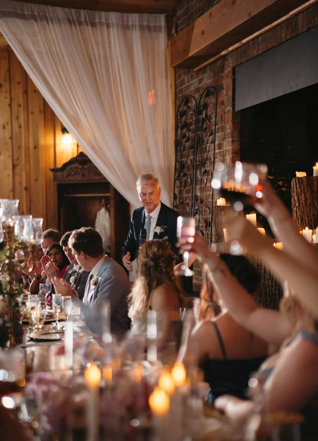 Father of the bride giving a toast as guests raise glasses during a rustic wedding reception dinner.