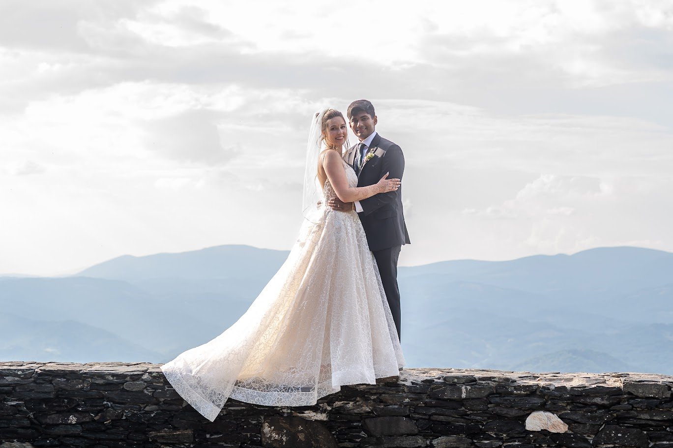 Bride and groom pose on a stone overlook with mountains behind them, the bride’s veil and skirt spread out.