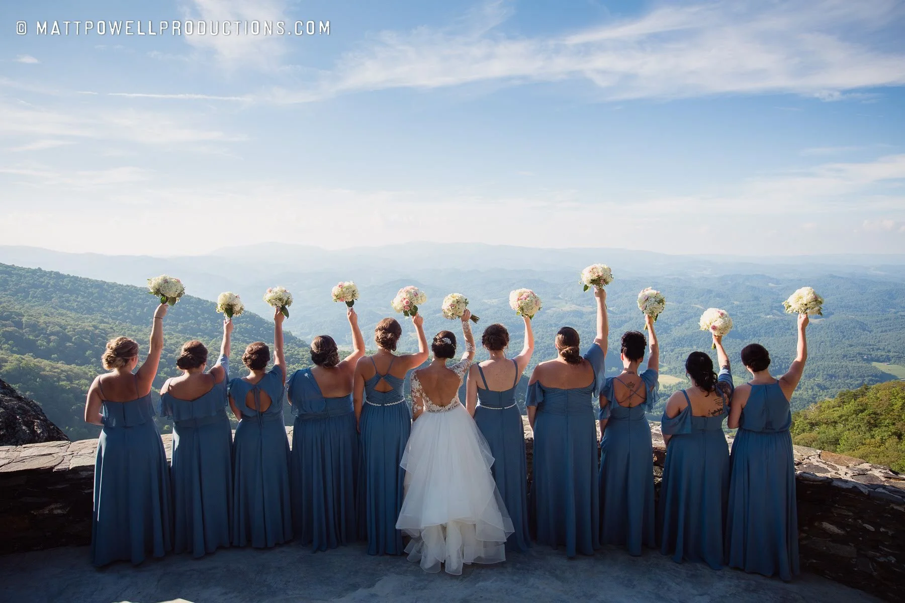 Bride and bridesmaids in blue dresses holding bouquets with a mountain view behind them.