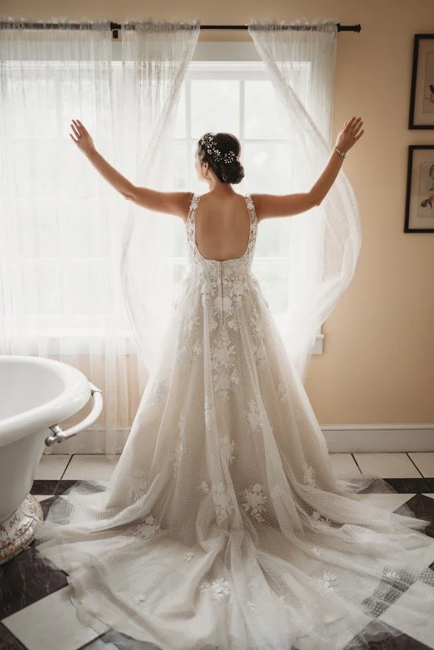 Bride in a strapless wedding dress standing with arms raised in soft window light before the ceremony.