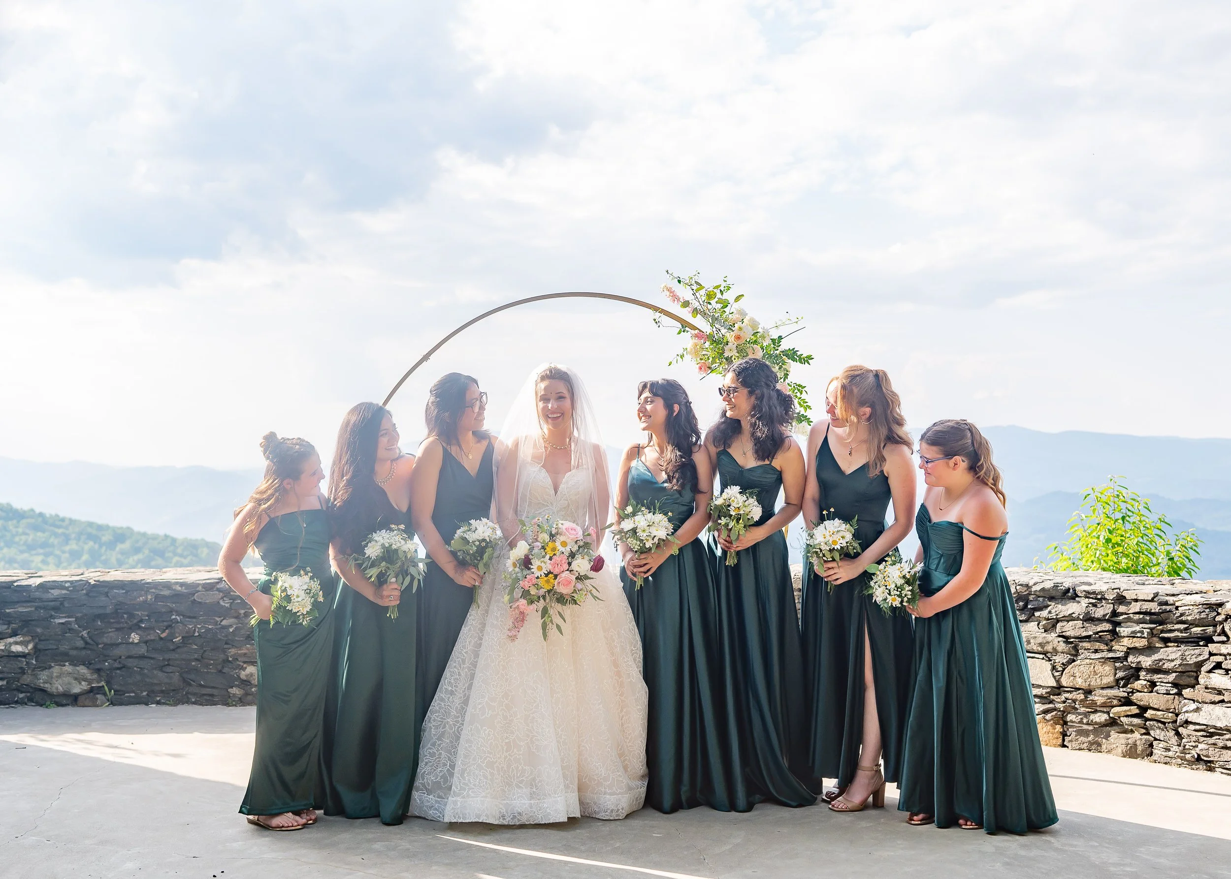 Bride posing with bridesmaids in emerald green dresses at a bright mountain overlook ceremony site.