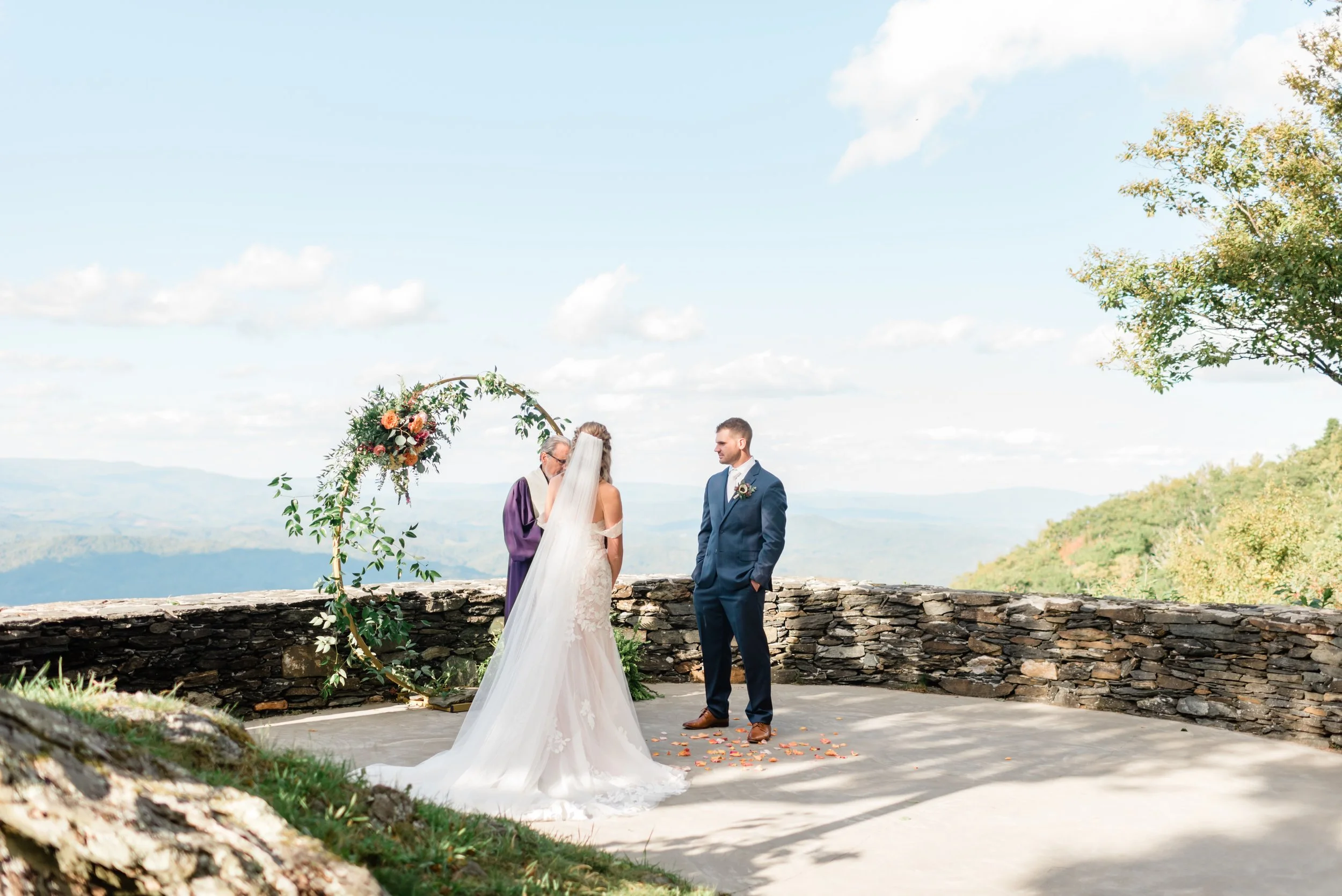 Mountain overlook wedding ceremony with the couple standing by a circular floral arch and stone wall.