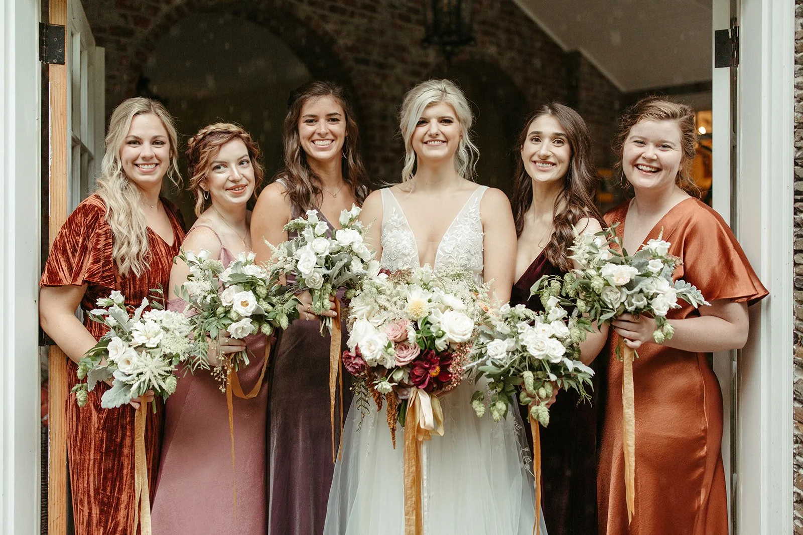Bride posing with bridesmaids in warm-toned dresses while holding lush white and burgundy bouquets.