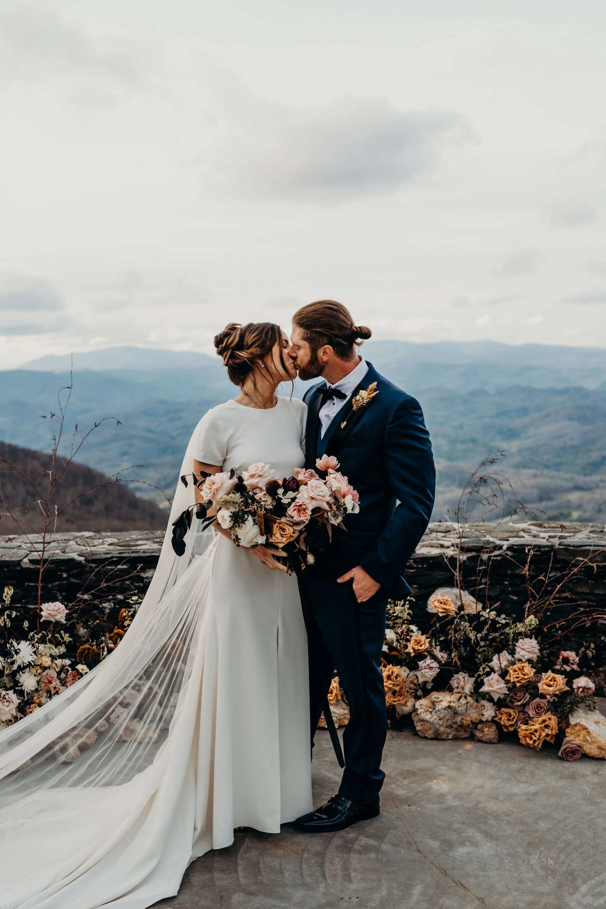 Bride and groom kissing at a mountain overlook with floral arrangements along a stone wall.
