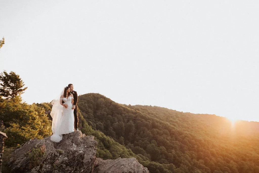 Wide shot of a bride and groom standing on a rocky overlook with the sun setting behind the mountains