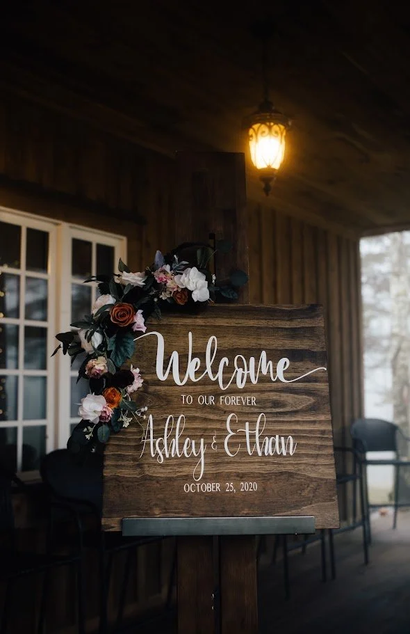 Rustic wooden wedding welcome sign with florals displayed on a porch under a warm lantern.