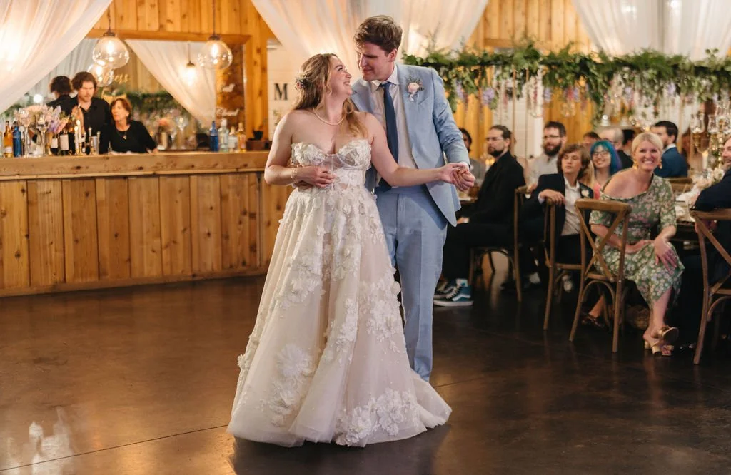Bride and groom smiling during their wedding reception entrance inside a rustic wood venue with guests seated nearby.