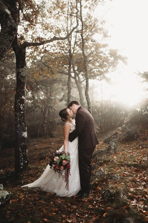 Bride and groom kissing during a misty woodland wedding portrait with autumn trees and a cascading bouquet.