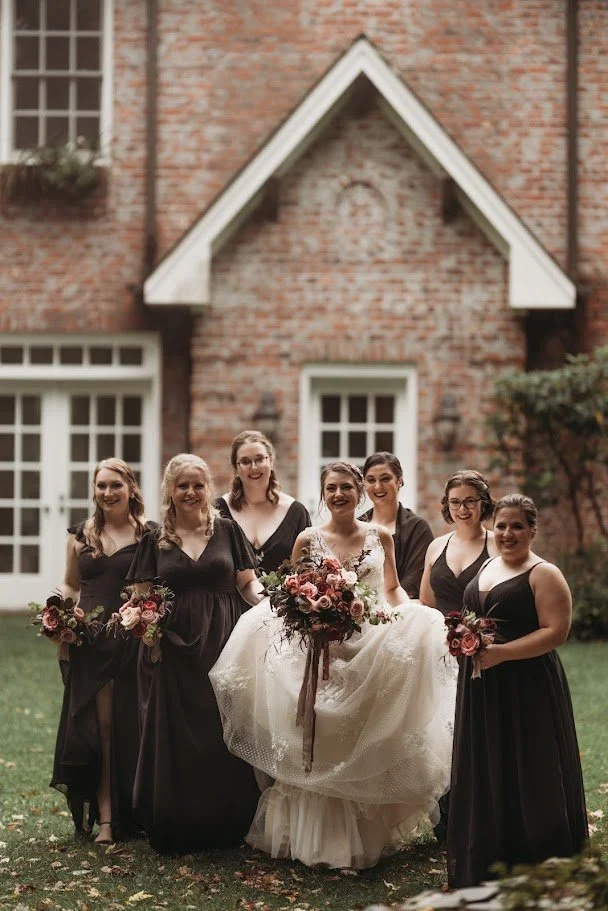 Bride posing with bridesmaids in black dresses holding autumn bouquets outside a brick estate wedding venue.