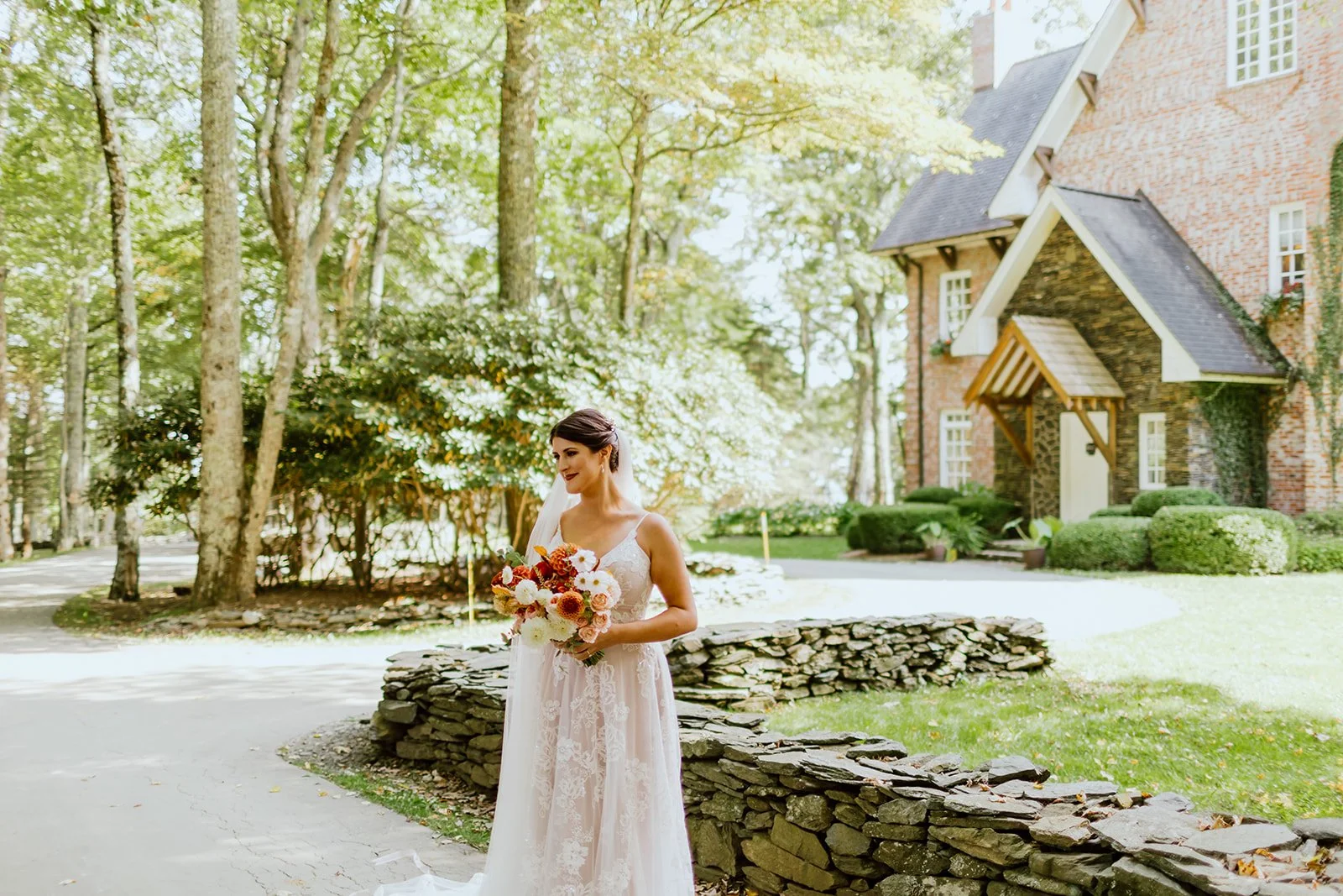 Bride holding an orange and white autumn bouquet outside a brick estate wedding venue with stone walls and trees.