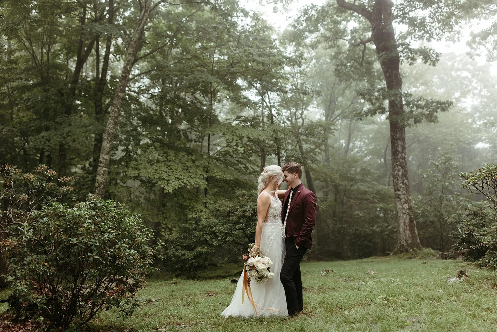 Bride and groom standing together in a misty forest wedding portrait with the groom wearing a burgundy suit.