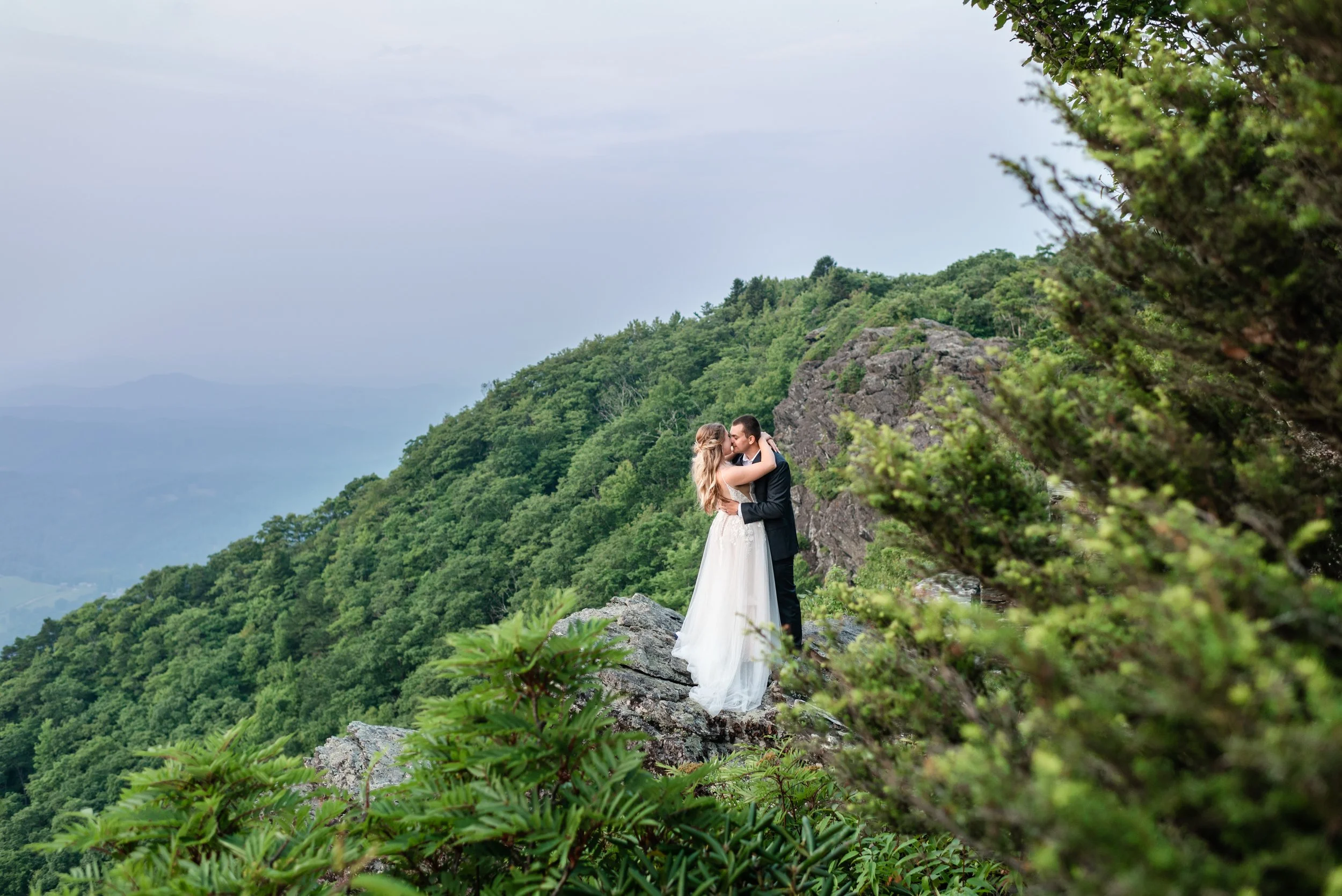 Bride and groom embracing on a rocky Blue Ridge mountain cliff surrounded by lush green forest and scenic mountain views.