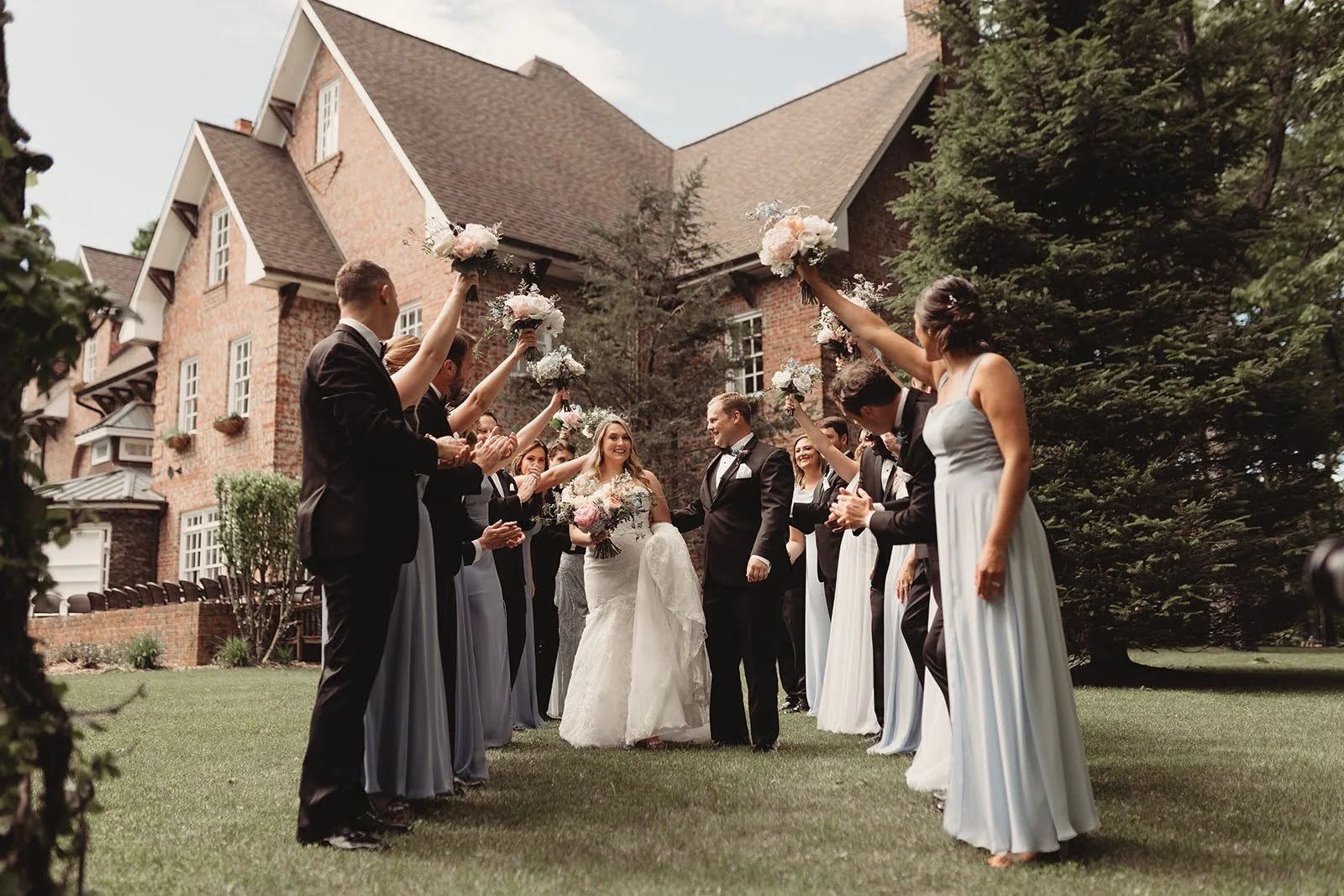 Wedding party forms a tunnel outside a venue, cheering as the couple walks through holding bouquets overhead.