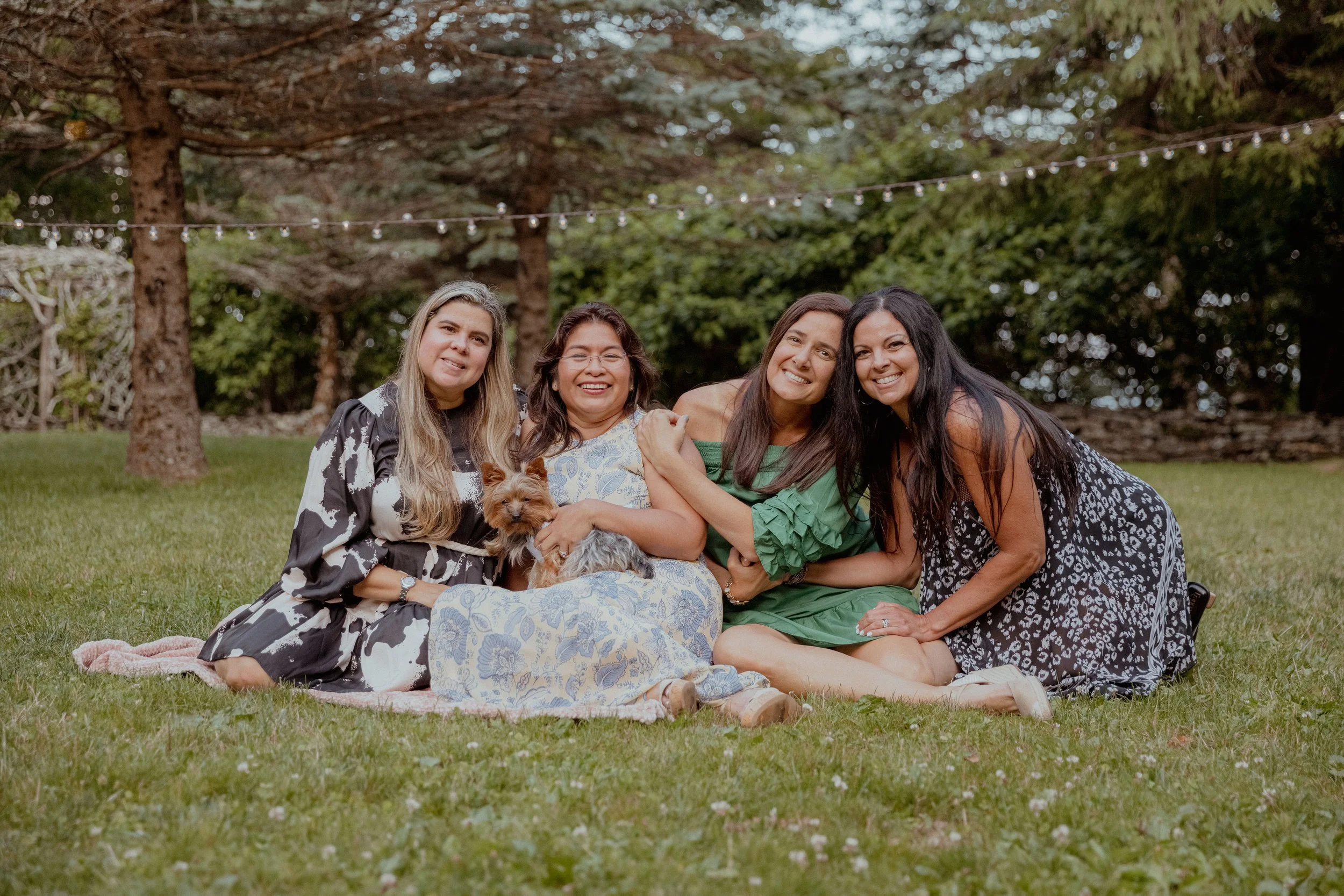 Four women sitting together on a lawn during a casual wedding weekend event with string lights in the background.