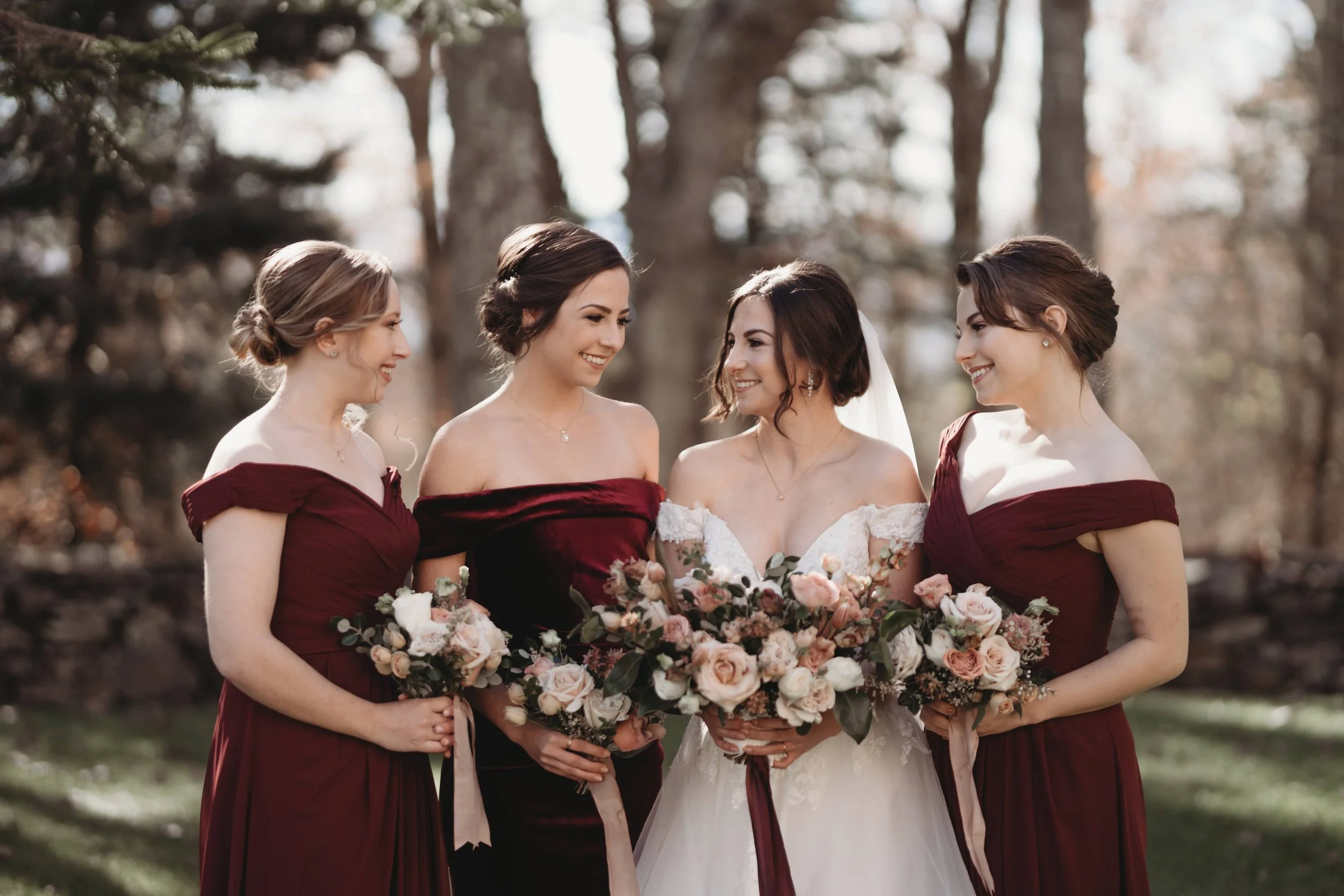 Bride with bridesmaids in burgundy dresses holding bouquets during outdoor portraits.