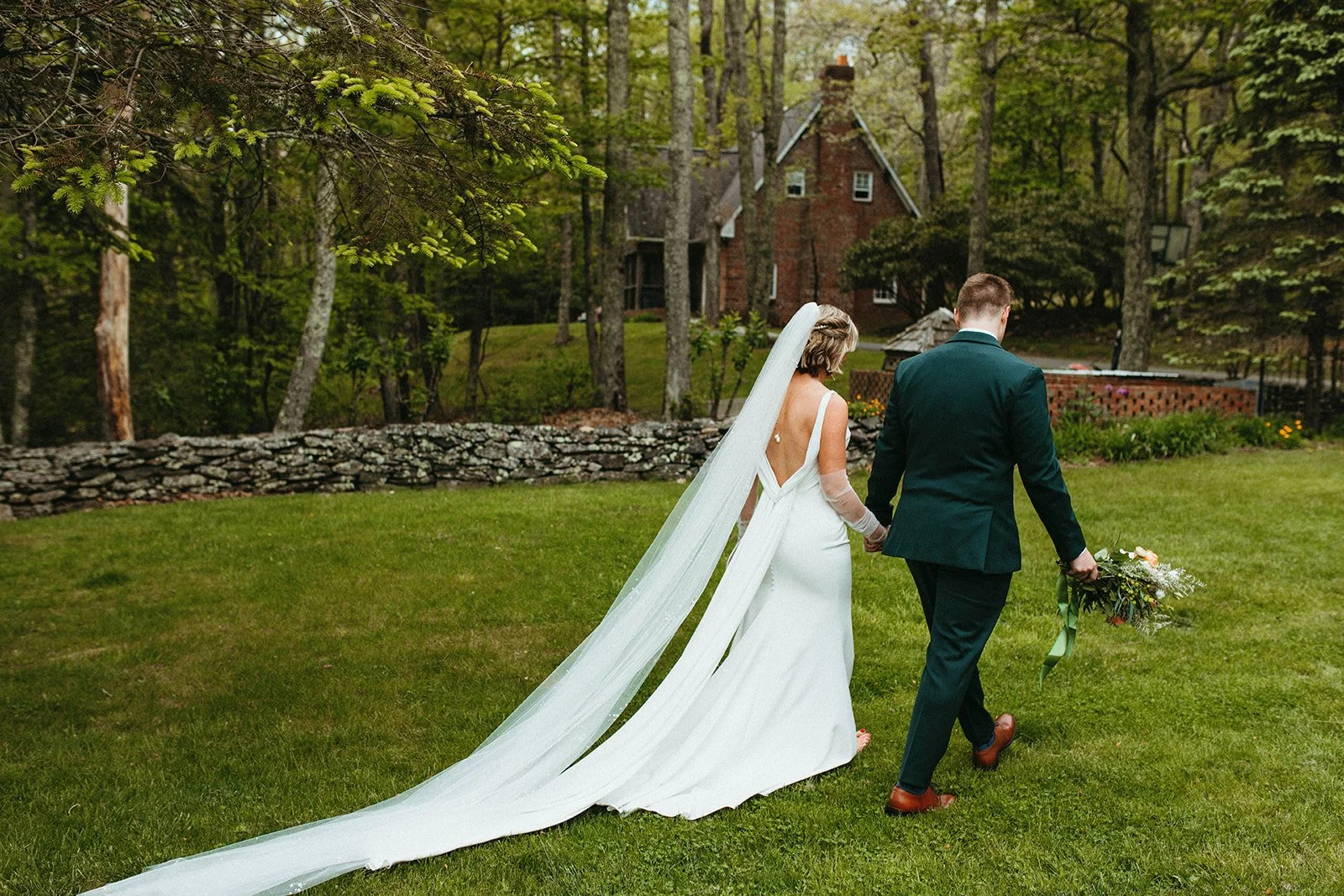 Bride and groom walk across a grassy lawn with the bride’s long veil trailing behind near a brick building.