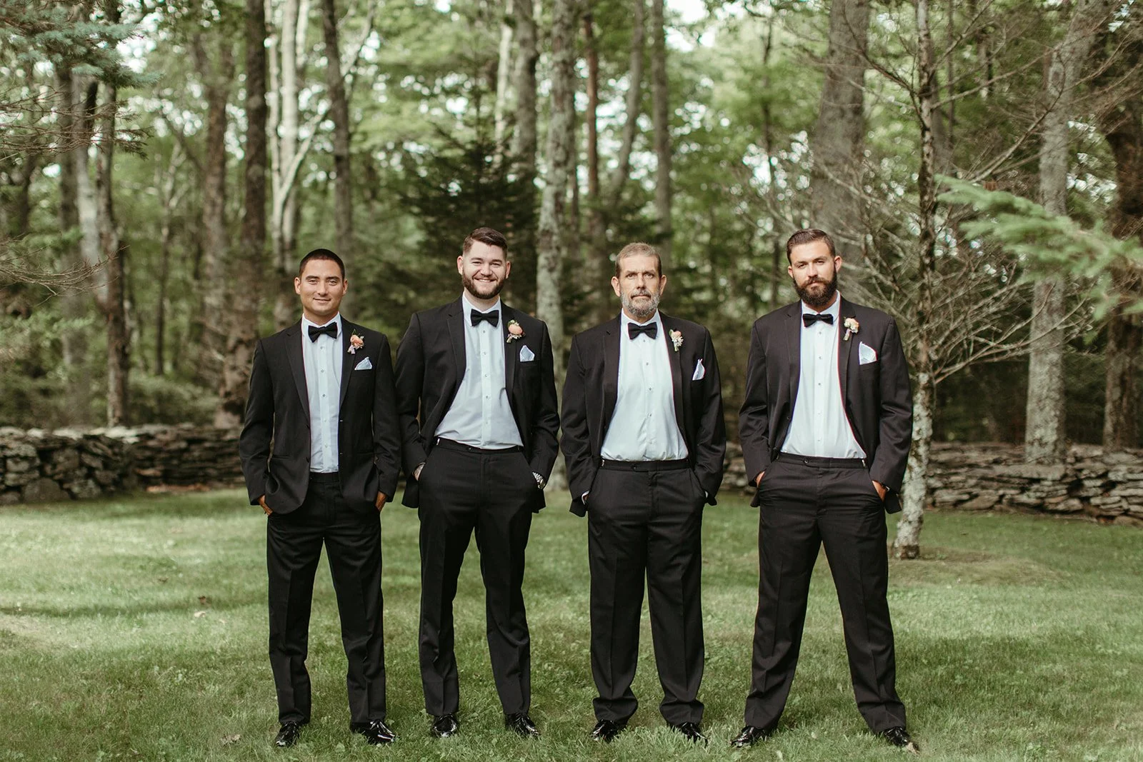 Groomsmen standing in black suits and bow ties during outdoor wedding portraits in the woods.