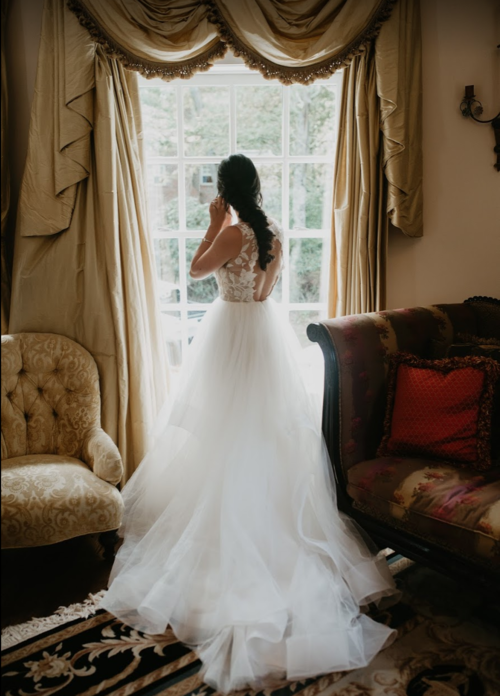 Bride in a lace wedding gown standing by a large window in a softly lit room