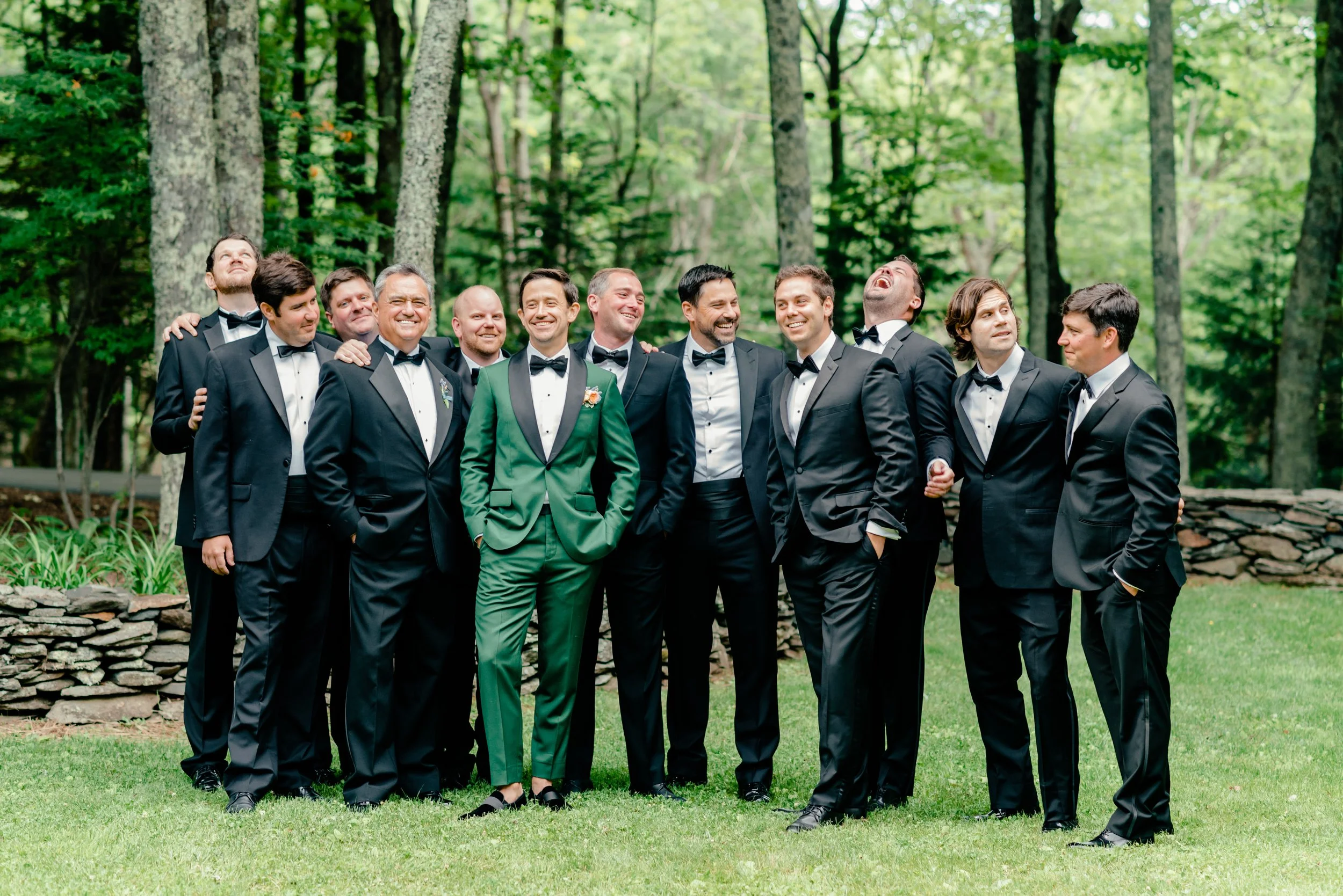 Groom in a green suit laughing with groomsmen in black tuxedos during outdoor wedding portraits.