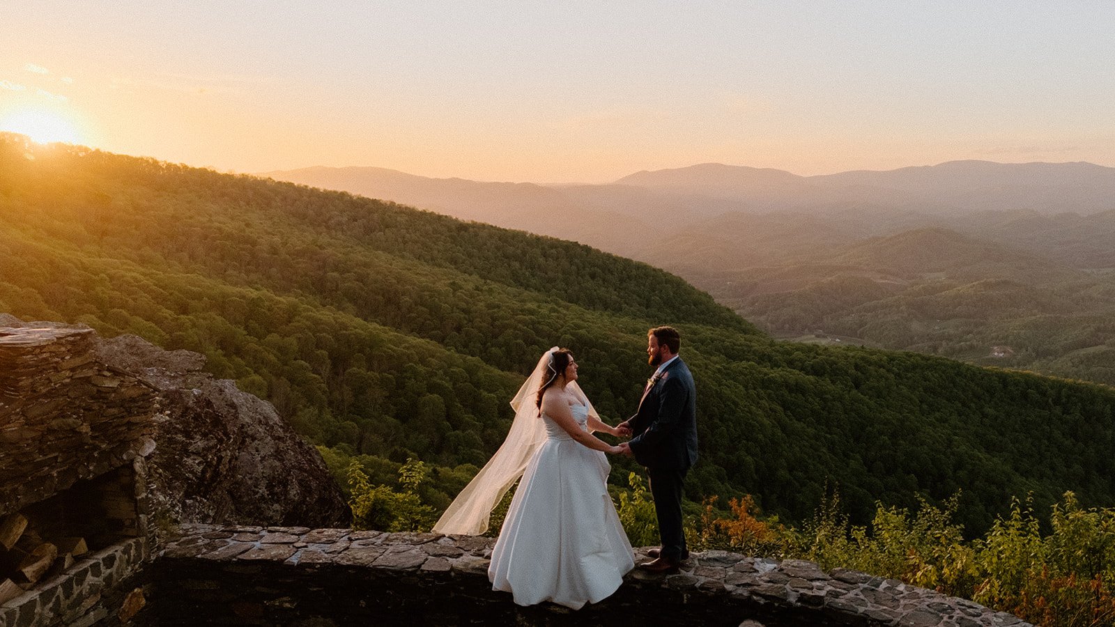 Bride and groom holding hands at sunset on a mountain overlook with sweeping views.