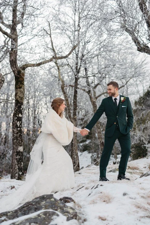 Winter wedding couple holding hands in snowy woods among bare trees.