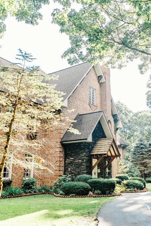 Close view of a brick wedding venue entrance with timber awning, stone accents, and manicured shrubs along the driveway.