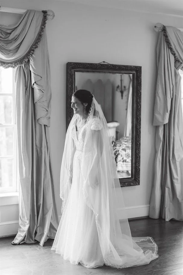 Black-and-white portrait of a bride in her veil standing by a window and mirror while getting ready