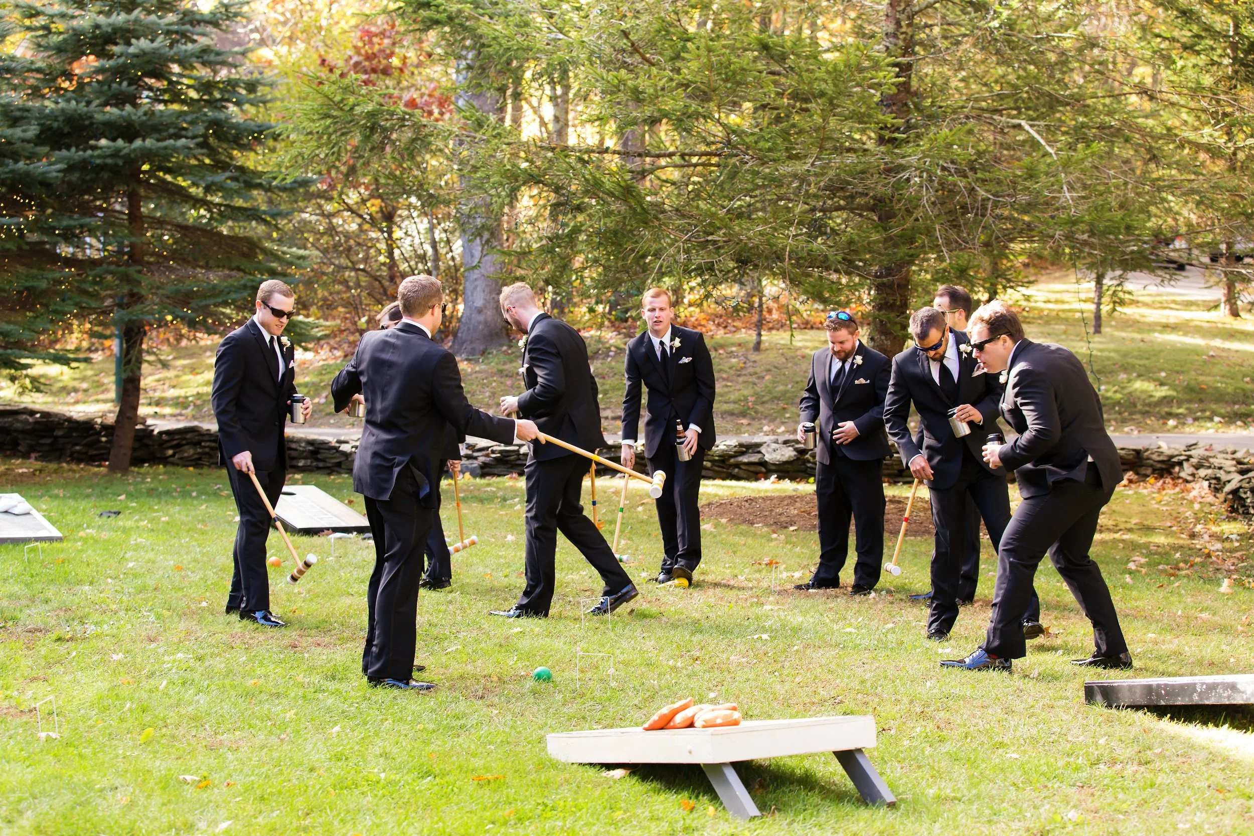 Groomsmen in navy suits playing lawn games outdoors during a wedding weekend celebration surrounded by trees.