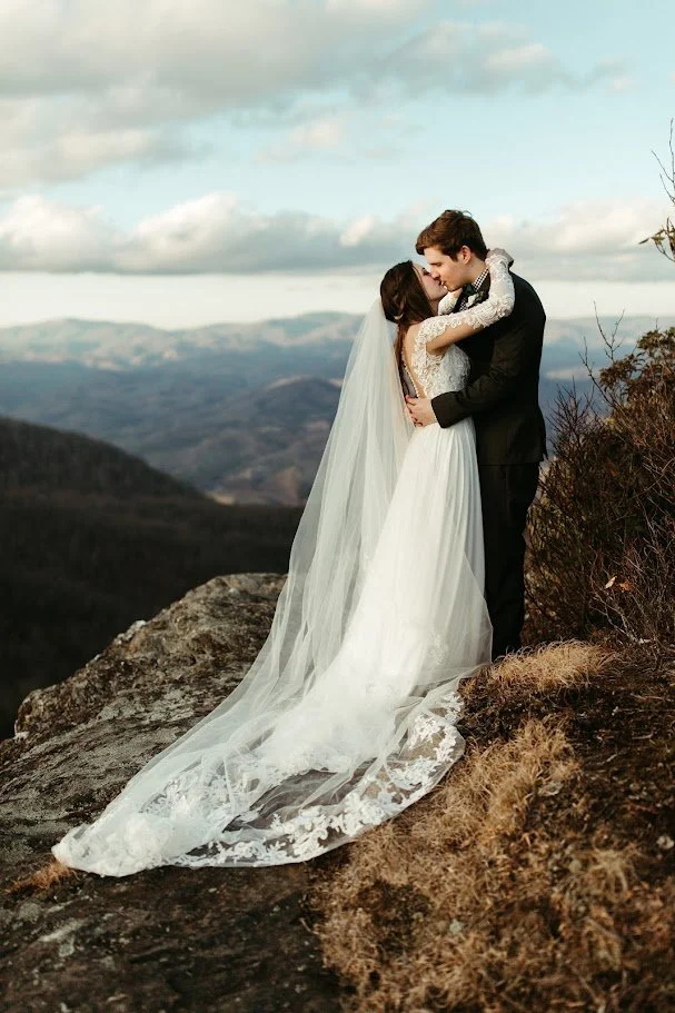 Bride and groom embracing on a rocky mountain overlook with layered mountain views behind them.