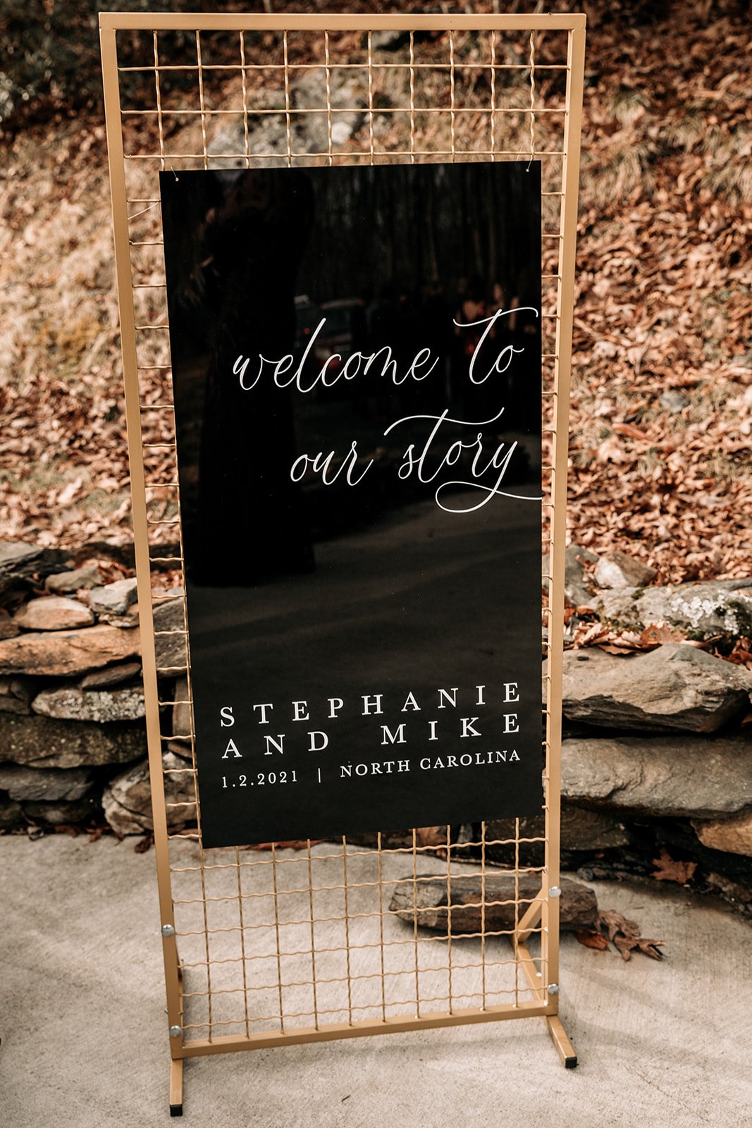 Black welcome to our story wedding sign displayed outdoors in a wooded ceremony setting.