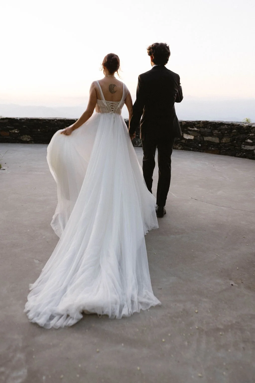 Bride and groom walking hand in hand toward a stone terrace with the bride’s long white train flowing behind her.