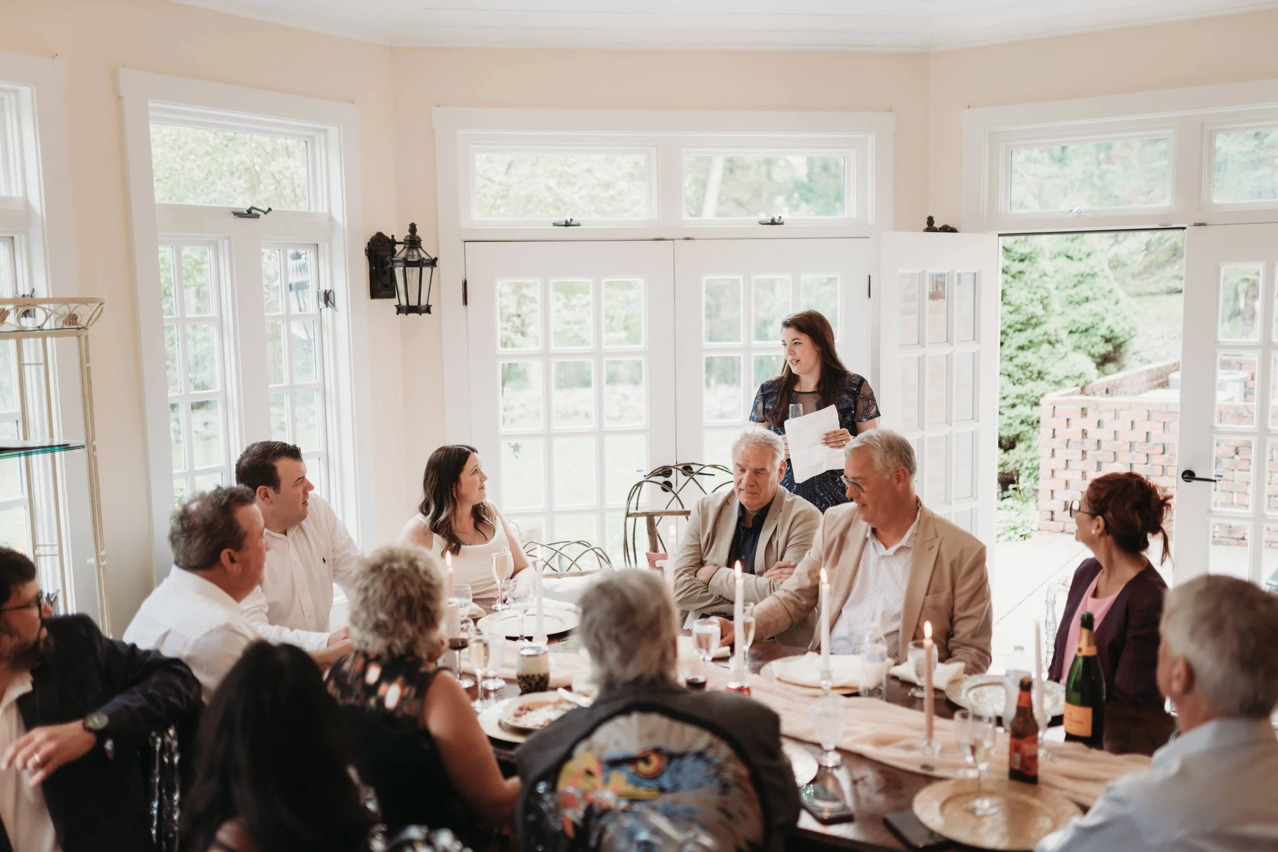 Guest giving a toast during an intimate indoor rehearsal dinner in a bright estate room with candles, wine, and seated guests.
