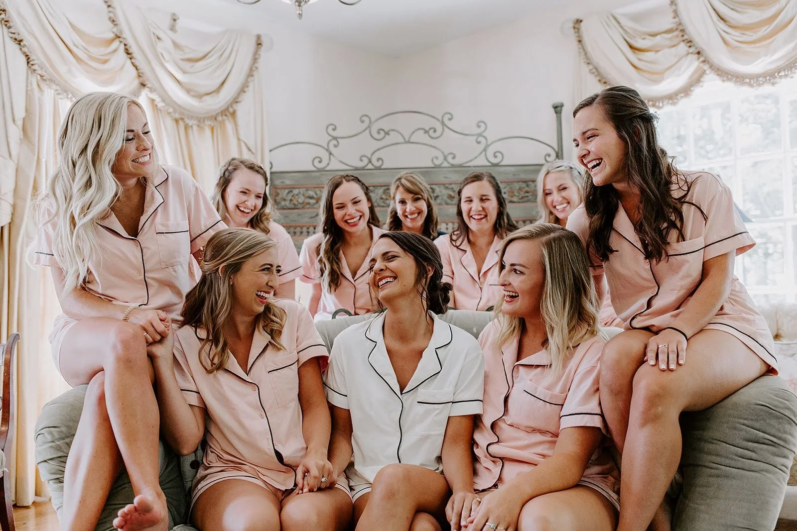 Bride and bridesmaids laughing together in pink pajamas while getting ready in an elegant bridal suite.