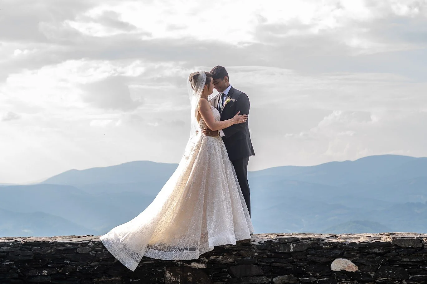 Bride and groom kiss on a stone overlook at sunset, her long veil flowing, with layered mountains in the distance.