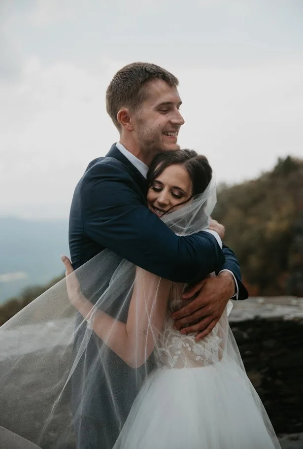 Bride and groom embracing at a mountain overlook with the bride’s veil wrapped softly around them.