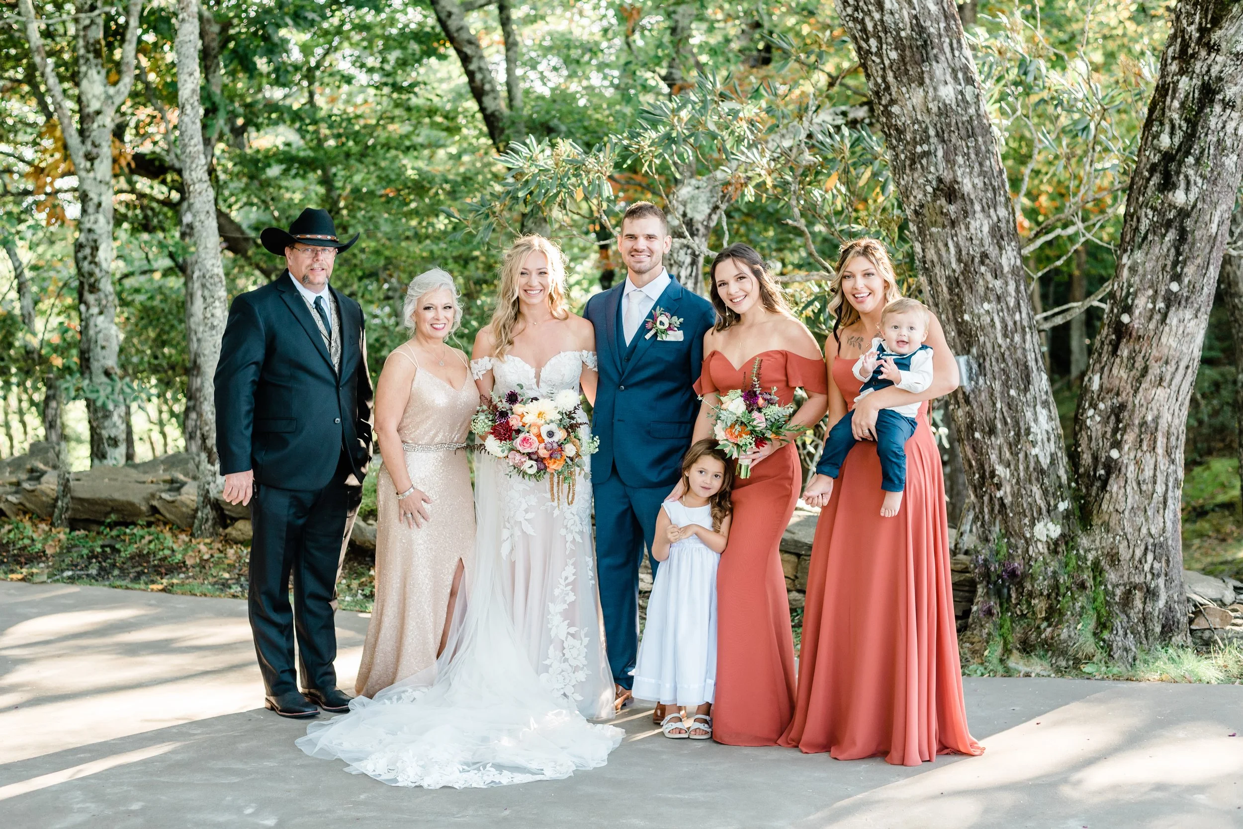 Bride and groom posing with family members outdoors in a wooded setting with autumn-colored dresses and floral bouquets.