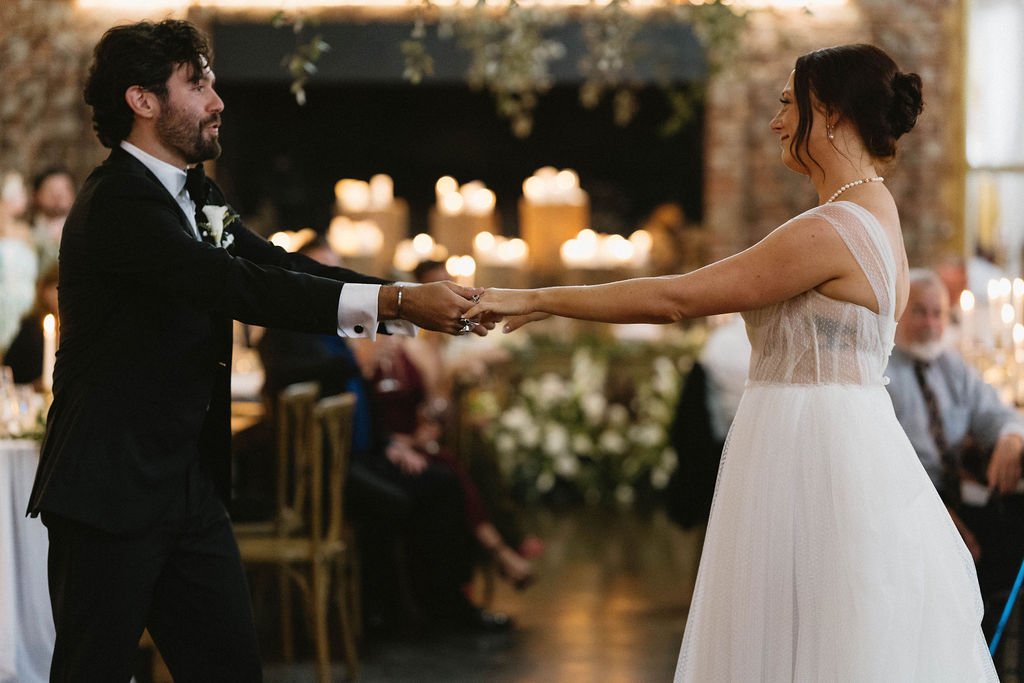 Bride and groom holding hands during their first dance in a candlelit reception room with brick walls and floral decor.