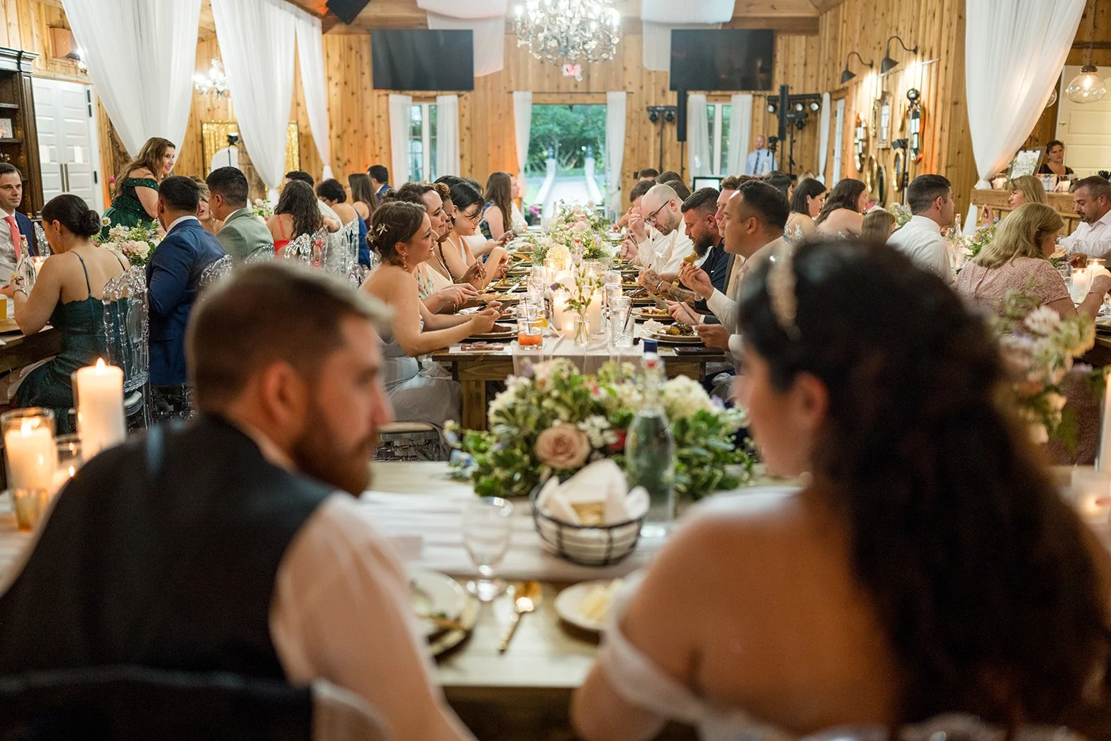 Guests seated at long wedding reception tables with candles, greenery, and soft floral centerpieces in a rustic venue.