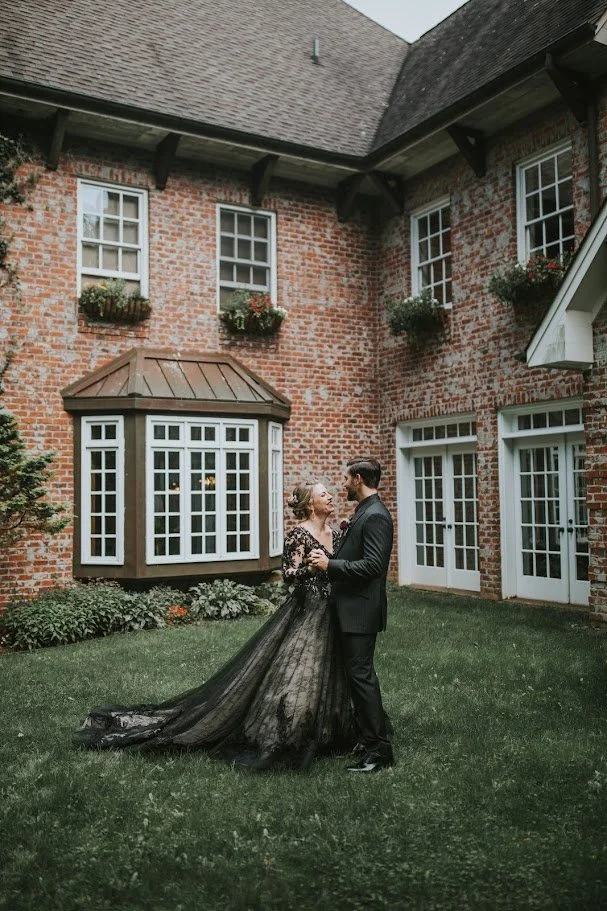 Couple standing in a courtyard in front of a brick building, bride wearing a dramatic black wedding gown.