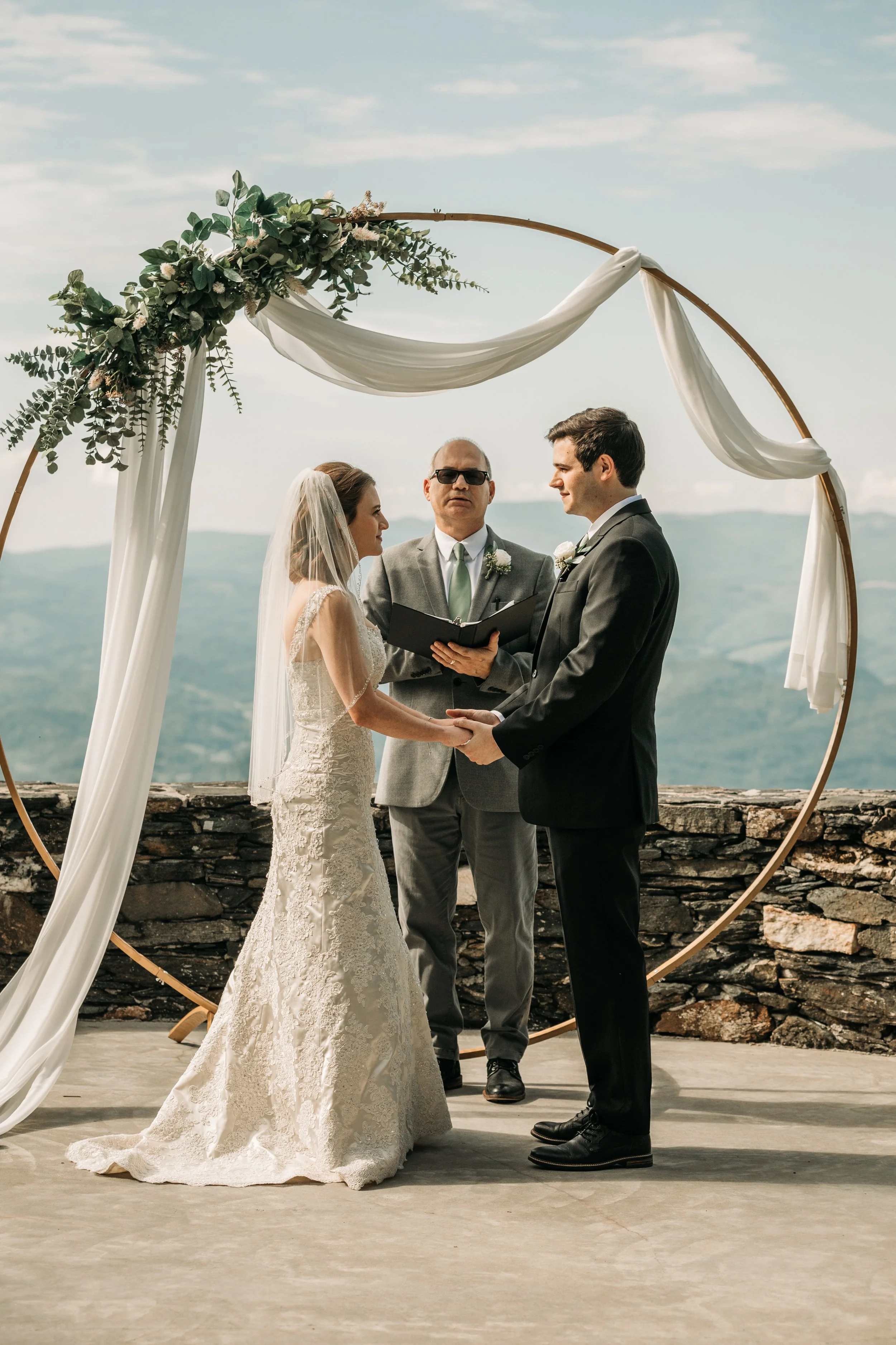 Bride and groom holding hands during a mountain overlook wedding ceremony beneath a white draped arch.