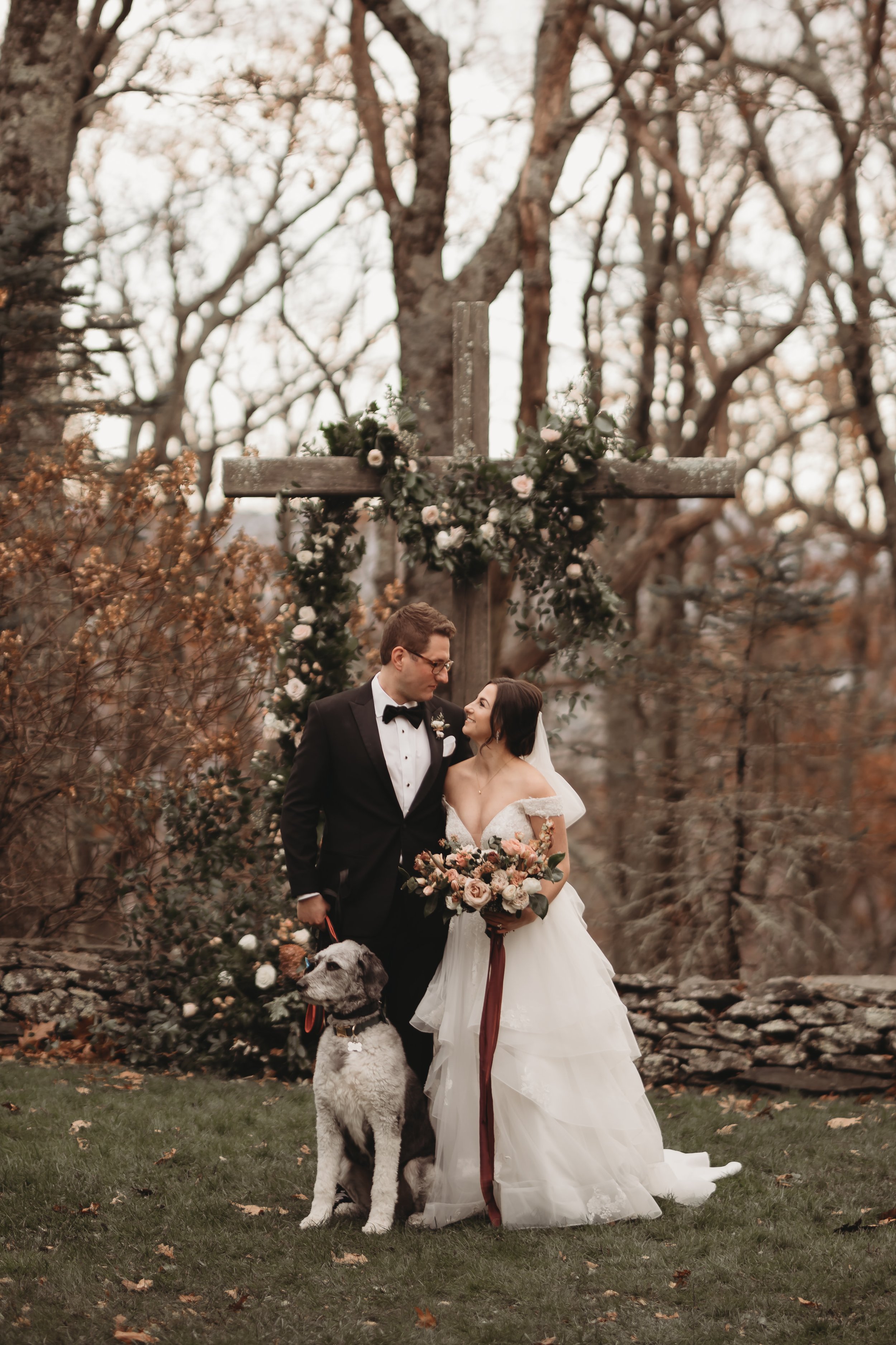 Bride and groom posing with their dog during an outdoor estate wedding, with a floral arch, stone wall, and autumn trees behind them.